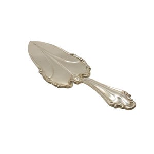 May include: Silver-colored pie server with a decorative handle and a leaf-shaped blade. The blade has a central vein design and ornate edging. The handle curves gracefully, ending in a decorative flourish.