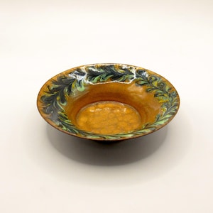 Puede incluir: Un cuenco redondo de cerámica con un interior naranja texturizado. El borde está decorado con un patrón de hojas verdes, negras y amarillas. El cuenco está sobre un fondo blanco.