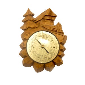 May include: A wooden weather barometer with a carved chalet and tree design. The barometer features a brass-colored dial with a needle indicating the weather conditions. The words "STURM", "REGEN", "VERANDERLICH", "SCHON", and "TROCKEN" are visible on the dial.