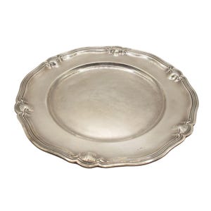 Puede incluir: Un plato de color plateado con borde festoneado y detalles ornamentados. El plato tiene un centro ligeramente cóncavo y un acabado pulido, adecuado para servir alimentos. Los elementos decorativos están espaciados uniformemente alrededor del borde.