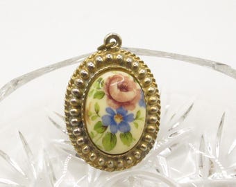 Puede incluir: Colgante ovalado con diseño floral. El colgante presenta una rosa pintada y flores azules sobre un fondo crema, enmarcado por un borde dorado con cuentas. Un pequeño lazo en la parte superior para sujetar a una cadena.