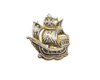 May include: A gold and black metal brooch shaped like a sailing ship. The ship features detailed sails, one of which displays a cross emblem, and a hull with decorative patterns. The design includes a mast and rigging, rendered in a metallic finish.