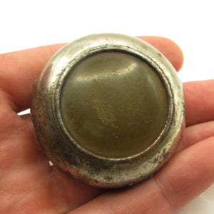 May include: A circular, vintage metal button with a worn silver appearance. The button has a central, olive-green, slightly curved insert. The button is held in a hand, highlighting its size and texture.