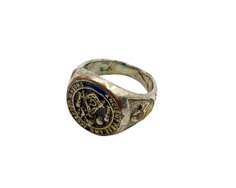 May include: A silver-toned ring with a detailed emblem. The ring features a blue and gold design with text around the edges. The ring has a worn appearance, suggesting age and use. The ring is likely a piece of jewelry.