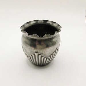 以下が含まれることがあります： 縁がギザギザで、底に装飾的なリブ模様が施された、小さな銀色の金属製のプランターです。