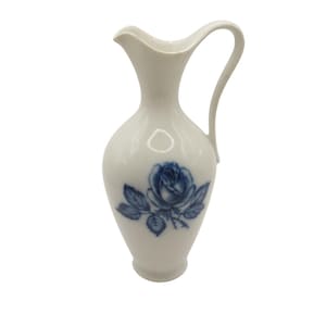 May include: A white ceramic pitcher with a blue rose design. The pitcher has a handle and a narrow neck.