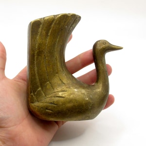 May include: A brass bird-shaped figurine with a weathered, antique finish. The object features a stylized, upright tail with etched feather details and a smooth, curved neck leading to a pointed beak. The piece has a solid, heavy appearance.