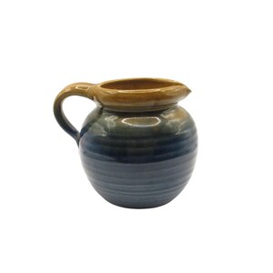 May include: A ceramic pitcher with a rounded body and a curved handle. The upper portion features a warm tan glaze, while the lower section transitions into a deep blue hue. The surface displays subtle horizontal ridges throughout the exterior.