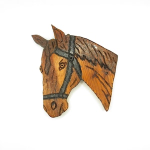May include: A wooden horse head carving, showing detailed wood grain and a dark bridle. The horse is brown with darker shading, and the bridle is black. The carving is set against a white background.
