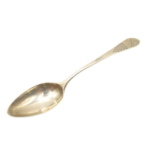 Puede incluir: Una cuchara de plata con un diseño ornamentado en el mango. La cuchara tiene forma ovalada y un acabado pulido.
