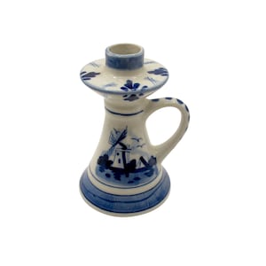 Può includere: Un portacandele in ceramica bianca e blu con un disegno di mulino a vento. Il portacandele presenta un manico e una base circolare. Il design include accenti floreali blu e una scena di mulino a vento, tipica della ceramica di Delft.
