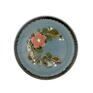 Könnte beinhalten: Ein runder Keramikteller mit hellblauer Glasur. Der Teller hat ein Blumendesign mit einer rosa Blume, grünen Blättern und weißen Akzenten. Der Rand ist dunkelgrau.