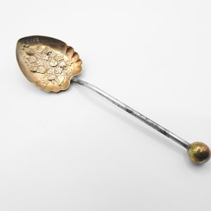 Pode incluir: Uma colher de prata e ouro com um design floral decorativo na tigela e uma bola redonda na ponta do cabo.