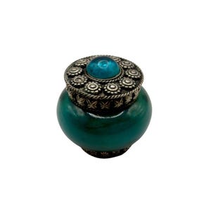 Può includere: Un piccolo barattolo color verde acqua con coperchio argentato. Il coperchio è ornato da una pietra turchese centrale e da un motivo floreale decorativo. Il barattolo ha una forma arrotondata.
