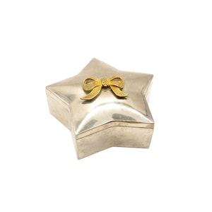 Può includere: Una scatola argentata a forma di stella con un fiocco dorato sul coperchio. La scatola ha un coperchio incernierato e una finitura lucida. Il fiocco dorato è un elemento decorativo.