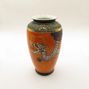 Può includere: Un vaso in ceramica arancione con un dettagliato disegno di drago bianco. Il vaso ha una base e un bordo neri, con accenti dorati e verdi vicino alla parte superiore. Il drago è finemente disegnato, aggiungendo un tocco artistico di ispirazione asiatica.
