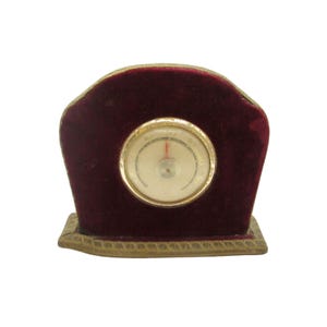 May include: A vintage thermometer with a gold-colored frame and a white face with black markings. The thermometer is set in a burgundy velvet base with a gold-colored trim.