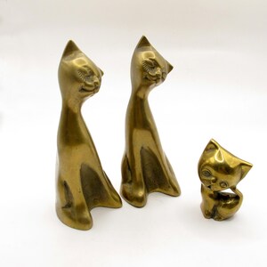 May include: Three brass cat figurines in varying sizes. Two tall, slender cats stand with elongated necks and closed eyes, while one smaller, compact cat sits with its head tilted and wide eyes. Each piece features a polished metallic gold finish.