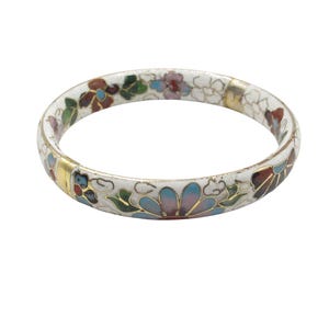Può includere: Un braccialetto a cerchio in smalto bianco con un motivo floreale. Il braccialetto presenta fiori colorati nelle tonalità di blu, rosso, verde e rosa. Accenti dorati sono visibili sulla superficie del braccialetto. È un gioiello.