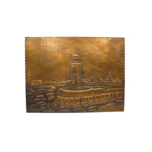 May include: A bronze-coloured metal wall art piece showing a coastal cityscape. The artwork features a lighthouse, buildings, and water, with a textured surface and a decorative edge.