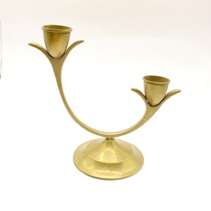May include: A brass dual-candle holder featuring a curved, U-shaped central arm that connects two individual candle cups. Each cup is supported by three small, petal-like metal prongs. The entire structure rests on a circular, flat base.