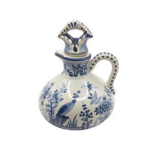 May include: A white ceramic jug with a blue floral and bird pattern. The jug features a curved handle and a decorative lid with a handle. The design includes flowers, leaves, and a bird perched on a branch.