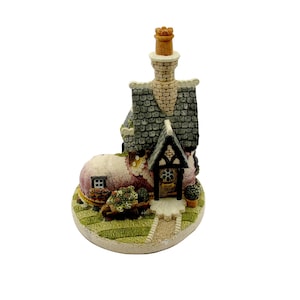 Op de afbeelding: Miniatuurbeeldje van een huisje met een rieten dak, een stenen schoorsteen en een kleine voordeur. Het huisje staat op een groene, grasachtige basis met een pad. Het beeldje heeft een grillige, sprookjesachtige esthetiek.