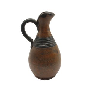 Puede incluir: Una jarra de cerámica marrón con un borde y un asa negros. La jarra tiene una superficie texturizada y una banda negra alrededor del medio.