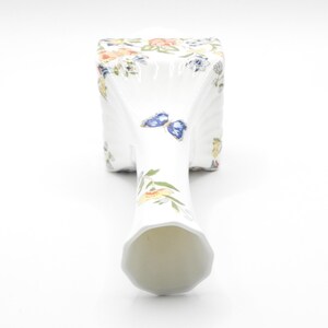 Puede incluir: Un jarr&oacute;n de porcelana blanca con base cuadrada y cuello alto y delgado. El jarr&oacute;n est&aacute; decorado con un patr&oacute;n floral y mariposas en tonos azules, amarillos y verdes.