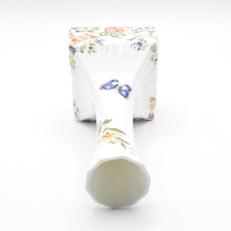 Puede incluir: Un jarr&oacute;n de porcelana blanca con base cuadrada y cuello alto y delgado. El jarr&oacute;n est&aacute; decorado con un patr&oacute;n floral y mariposas en tonos azules, amarillos y verdes.