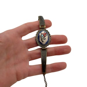 Könnte beinhalten: Ein Armband im Vintage-Stil mit einem ovalen Mittelstück mit floralem Design. Das Mittelstück hat einen weißen Hintergrund mit einer roten und blauen Blume. Das Armband hat einen bronzefarbenen Metallrahmen und eine Kette.