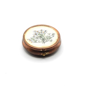 May include: A round, copper-colored pill box with a white lid featuring a floral design of white daisies with yellow centers.