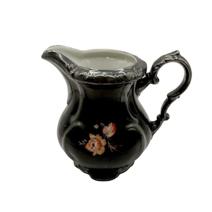 May include: A black porcelain creamer with a silver trim and floral design. The creamer has a rounded body and a handle with a decorative design.