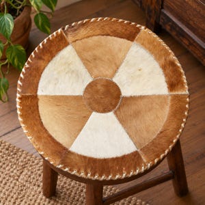 May include: A round stool featuring a seat upholstered in a patchwork of brown and cream cowhide leather. The circular seat is divided into alternating triangular segments with a central circular patch, all joined by visible white stitching.