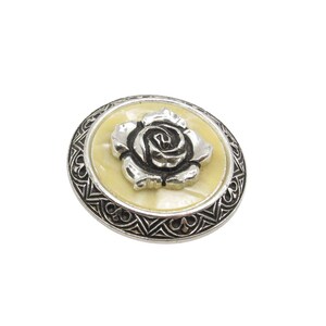 K&ouml;nnte beinhalten: Silberfarbene Brosche mit Rosenmotiv. Die Rose befindet sich auf einem cremefarbenen Hintergrund, umgeben von einem dekorativen Silberrand. Die runde Brosche hat einen verzierten, Vintage-Stil.