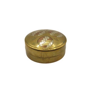 May include: A small, round brass box with a hinged lid. The lid features three circular inlays of woven metal in silver, copper, and brass. The box has a simple, classic design and a polished finish. The box is likely used for storing small items.