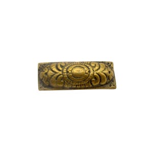 May include: A rectangular, antique brass drawer pull with an ornate, symmetrical design. The pull features a central oval shape surrounded by swirling floral and leaf patterns. The brass has a slightly aged, tarnished appearance.