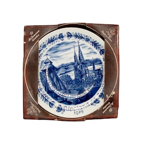 May include: A blue and white decorative plate featuring a detailed landscape scene of a church and surrounding structures. The plate is in a brown cardboard box with text that reads "Upland 1975". The plate has a floral border.