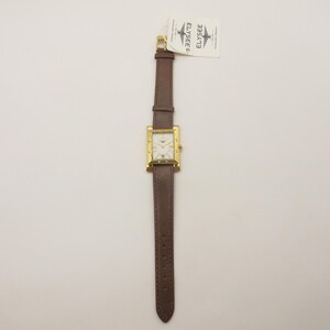 Puede incluir: Un reloj cuadrado dorado con esfera blanca y correa de cuero marrón. El reloj tiene una esfera rectangular con detalles dorados y una correa de cuero marrón. El reloj es nuevo y tiene una etiqueta.
