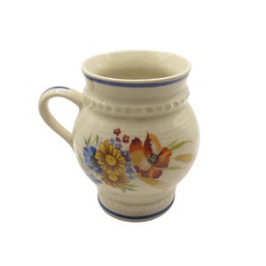 Può includere: Tazza in ceramica color crema con manico curvo e design floreale. La tazza ha un bordo e una base blu, con una fascia di piccoli punti in rilievo al centro. Il design floreale comprende fiori blu, gialli e arancioni.