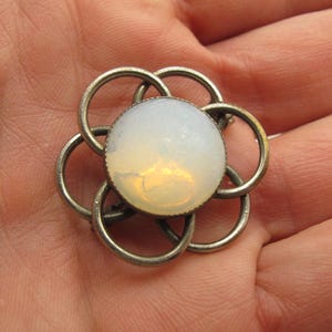 Op de afbeelding: Een ronde broche met een centrale, gladde, melkwitte cabochonsteen met subtiele iriserende accenten. De steen wordt omlijst door een reeks van zes in elkaar grijpende, zilverkleurige metalen ringen die een bloemachtig patroon vormen.