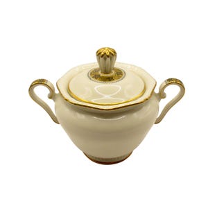 May include: Cream-coloured ceramic sugar bowl with a lid and two handles. The lid is topped with a gold-coloured finial. Gold trim accents the rim, handles, and lid. The sugar bowl has a scalloped edge.