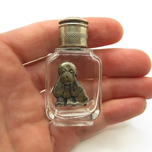 Puede incluir: Pequeño frasco de perfume de vidrio transparente con tapa de metal y una figura de perro de metal en el interior. El perro tiene orejas largas y caídas y una expresión triste.