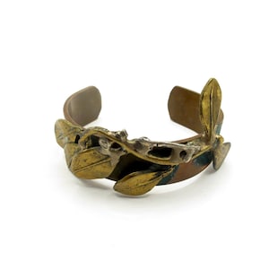 Pode incluir: Uma pulseira de punho com uma faixa de bronze e um design decorativo de folhas em tons de ouro e prata. As folhas são detalhadas e adicionam um elemento natural à joia.