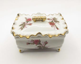 Puede incluir: Una caja de porcelana blanca con ribete dorado y diseño floral. La caja tiene una tapa con un asa dorada y se apoya en cuatro pequeñas patas.