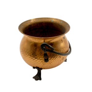 Può includere: Un piccolo calderone in rame martellato con un manico in metallo nero e tre piedini decorativi. Il vaso ha un corpo arrotondato e un bordo largo. Il rame ha una calda tonalità marrone-rossastra e il manico è fissato con rivetti.