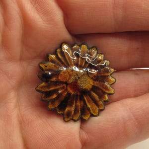 Può includere: Una spilla a forma di fiore, dettagliata e realizzata a mano. La spilla presenta un design a strati con petali marroni e dorati, un centro rosso e oro e una finitura lucida. La spilla è tenuta in una mano.