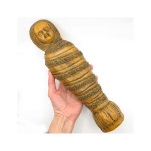 May include: A carved, tan-colored figurine resembling a mummy, held in a hand. The figure has a detailed face and is wrapped in bands with ornate designs. The bottom of the figure has a flared, fan-like shape. The figurine is approximately 10 inches long.