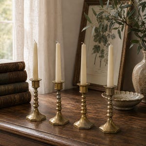 May include: Four matching brass candlesticks with a tiered, ribbed stem design and a hexagonal base. Each candlestick holds a single, slender, off-white taper candle. The metal has a muted, aged gold finish with visible signs of patina.