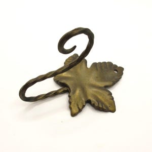 May include: A bronze-colored napkin ring in the shape of a leaf with a twisted stem and a curled top. The leaf has detailed veining and a hole near the edge. The napkin ring is made of metal.